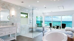 A modern bathroom with a glass-enclosed shower, a large bathtub, and a view of the ocean through large windows. - Luxury 6 Bedroom Villa on the Beach in Bodden Town (Breakers)