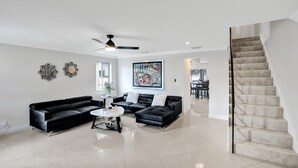 Ein geräumiges Wohnzimmer mit einer schwarzen Ledersitzgruppe, einem Couchtisch und einem Gemälde an der Wand. Eine mit Marmor ausgekleidete Treppe führt auf eine obere Ebene.