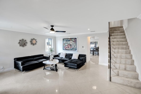 A spacious living room with a black leather sofa set, a coffee table, and a painting on the wall. A staircase with a marble finish leads to an upper level.