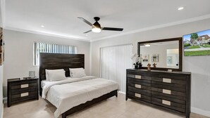 Ein Schlafzimmer mit einem Bett, zwei Nachttischen, einer Kommode, einem Deckenventilator, einem großen Spiegel und einem Fenster mit Jalousien.