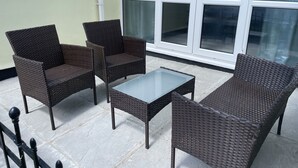 Een terras met rieten meubilair, waaronder twee stoelen en een bijzettafeltje, nabij een glazen deur.