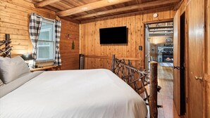 Un dormitorio de cabaña de madera con una cama, un televisor y una chimenea visible a través de una puerta abierta.