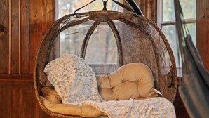 Una silla colgante de mimbre con un cojín y una manta, frente a una pared de madera con una ventana al fondo.