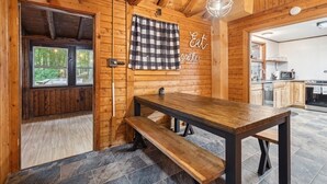 Interior de madera con una mesa de comedor y banco, un mantel a cuadros colgando, y un área de cocina visible a través de una puerta abierta.