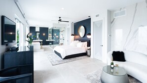 Ein modernes Schlafzimmer mit einem großen Bett, einem Fernseher, einem Duschbad mit Glastüren und einem weißen Sofa.