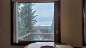 Ein Zimmer mit einem großen Fenster, das Blick auf einen Wald und Berge bietet. Auf dem Holztisch stehen eine Schüssel und Matten.