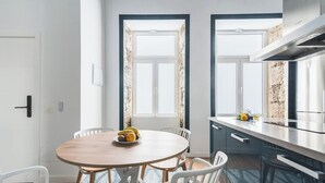A modern kitchen with a round wooden table, white chairs, and a fruit bowl. There are two windows with curtains, a stainless steel range hood, and a countertop with a vase of flowers.