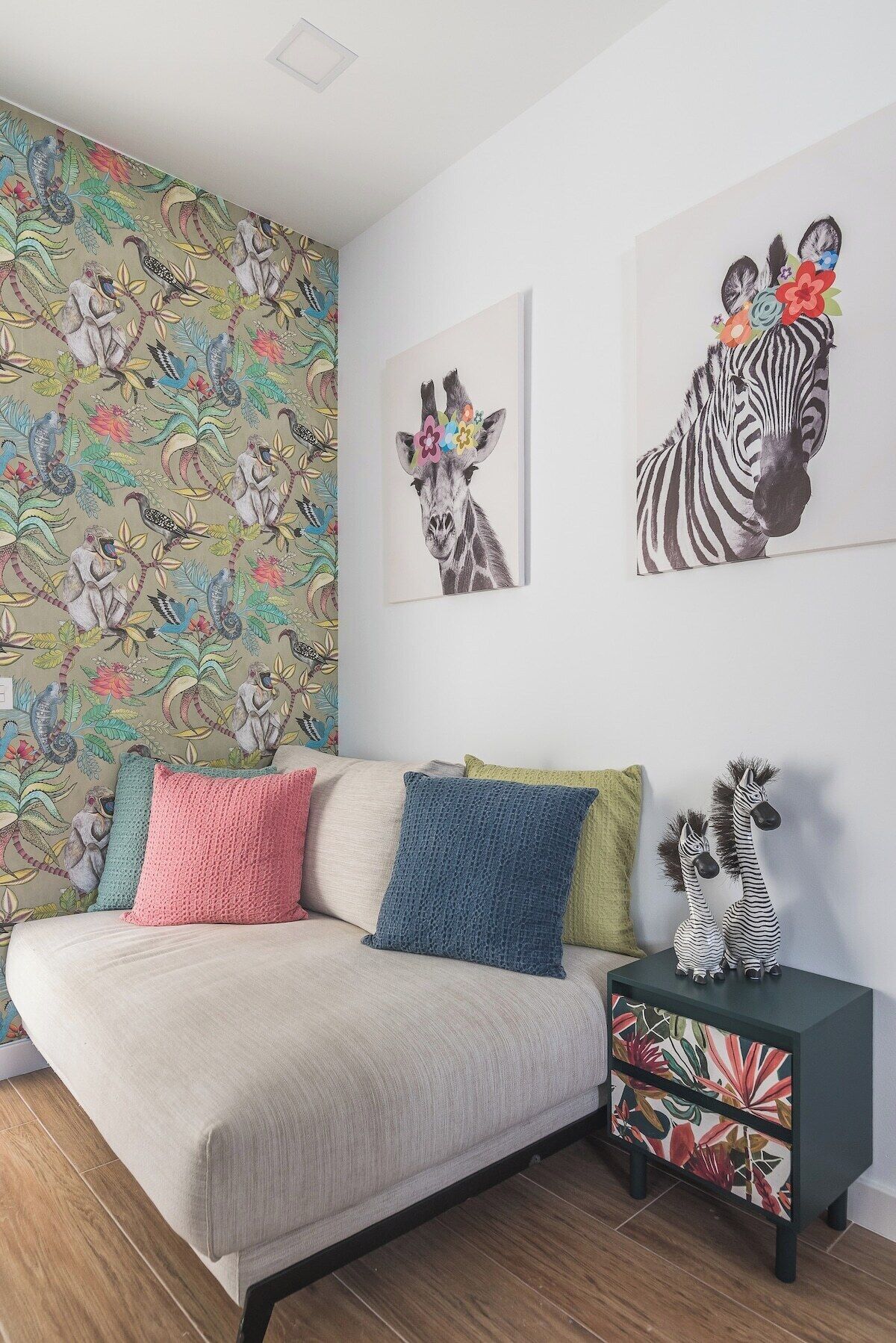 A bedroom with a sofa bed, colorful pillows, a side table with zebra figurines, and a wall with a floral wallpaper and two zebra paintings.