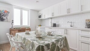 A modern kitchen with white cabinets, a microwave, and a coffee machine. There is a dining table with a green leaf-patterned tablecloth and white chairs.