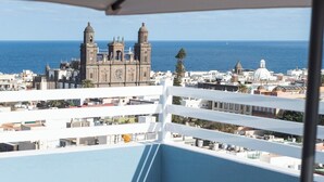 A balcony with a table, a chair, and a glass of wine, overlooking a coastal cityscape with historic buildings and a clear blue sky.