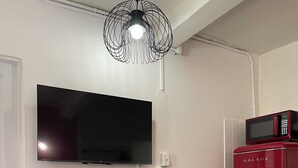 Ein kompakter Wohnbereich mit einem wandmontierten Flachbildfernseher, einem kleinen Esstisch mit zwei Stühlen, einem roten Kühlschrank und einer auffälligen schwarzen Pendelleuchte.
