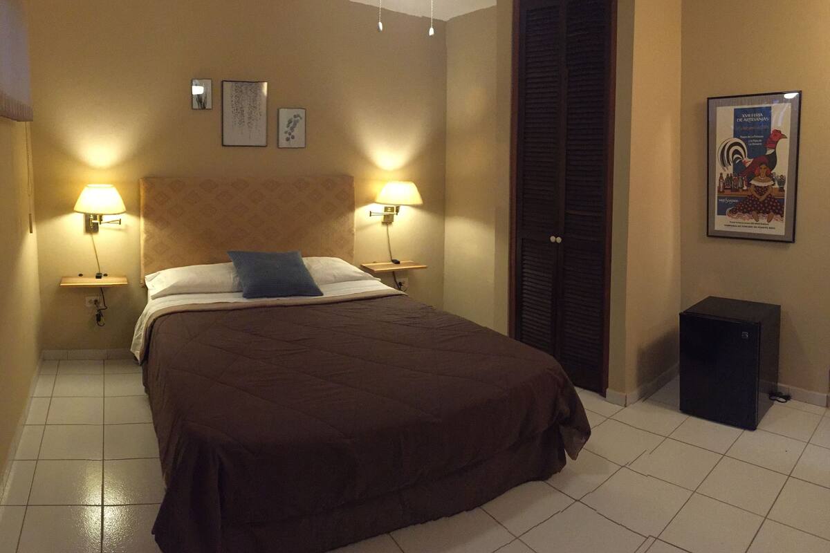 A hotel room with a large bed, two bedside lamps, a nightstand, a wardrobe, and a small black speaker.