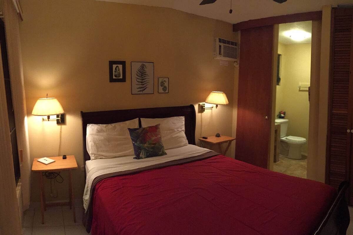 A hotel room with a bed, two bedside lamps, a red bedspread, and a wooden headboard.