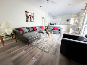 A modern living room with a grey sectional sofa, a coffee table, and a rug. There is a painting on the wall, a ceiling fan, and a view into the kitchen area.