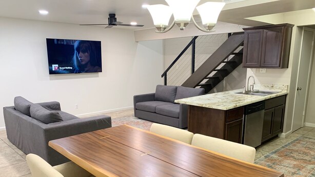 A modern kitchen with a dining area, grey sofas, a wooden dining table, and a TV mounted on the wall.