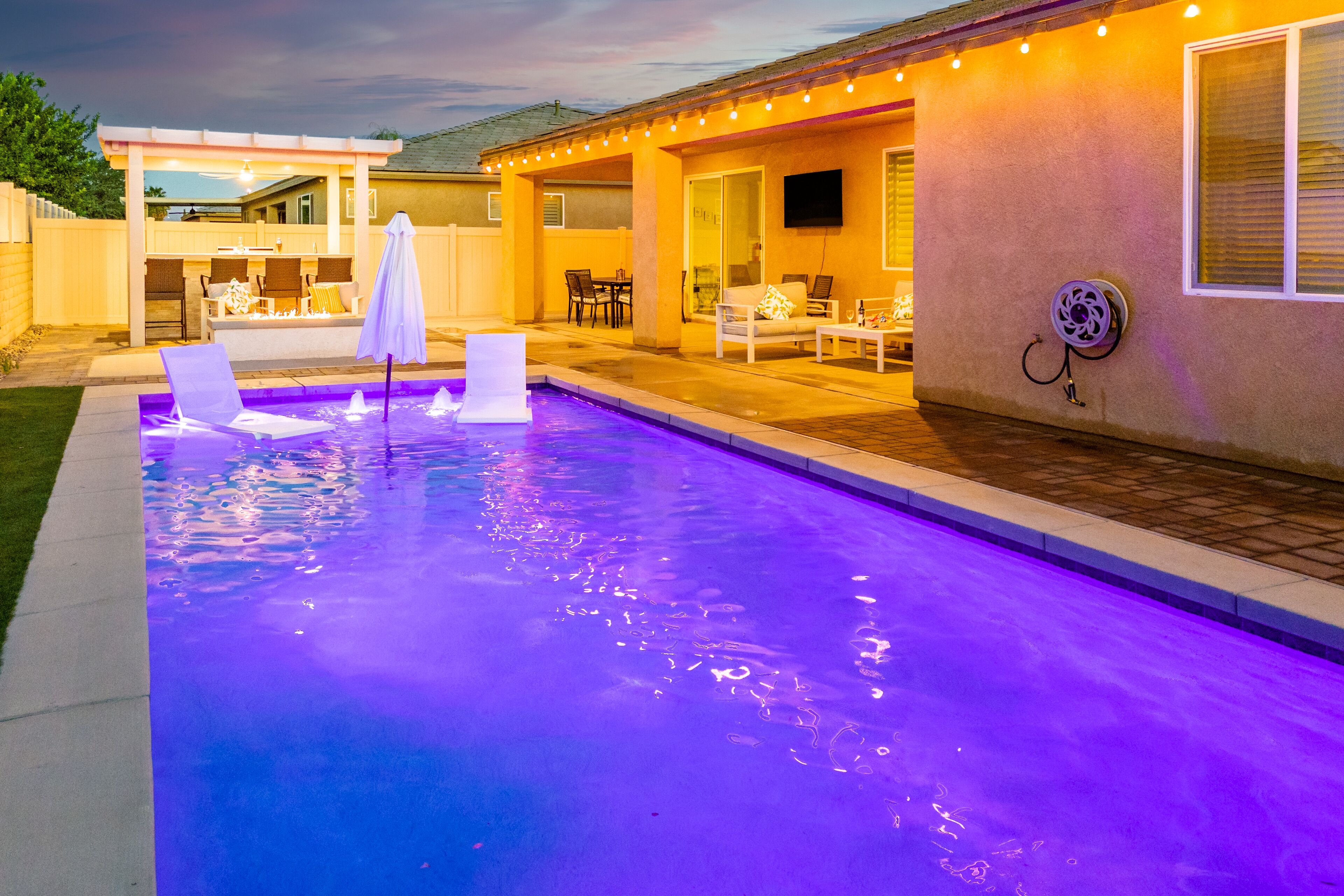 A swimming pool with a blue-lit edge, outdoor seating area, and a covered patio with string lights.