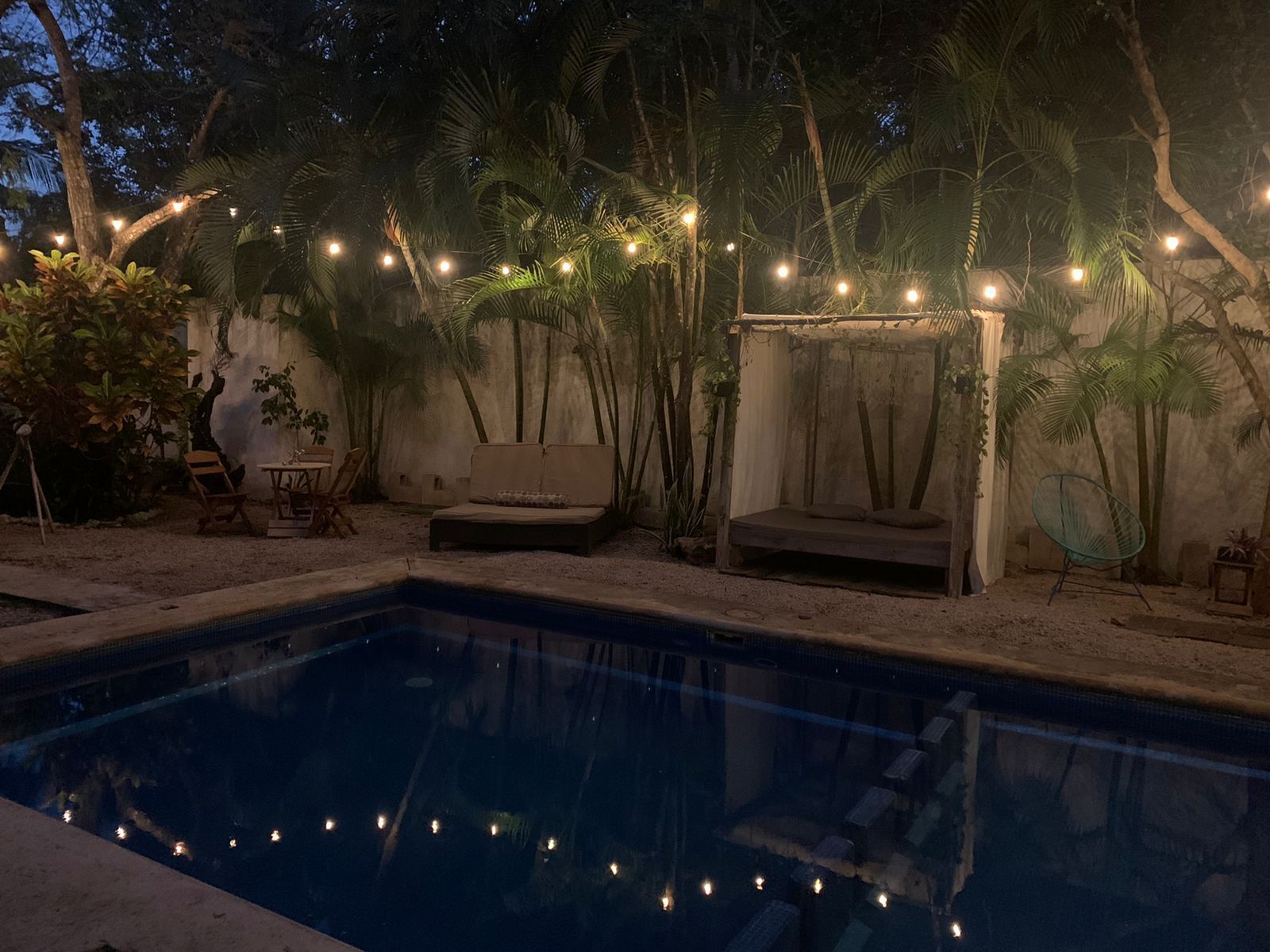 A pool area with a lounge chair, a small table, and a bench. There are palm trees and string lights around the pool.