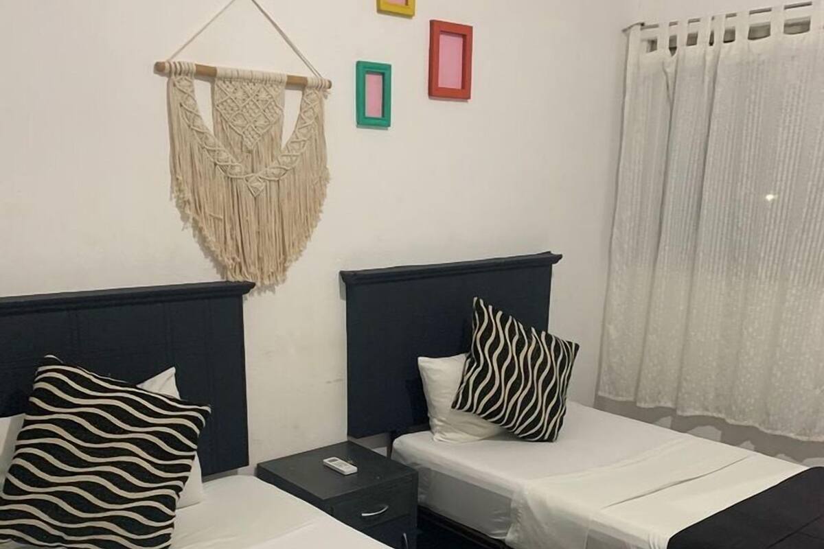 Hotellihuone, jossa on kaksi sänkyä. Jokaisessa sängyssä on mustavalkoraidallinen tyyny, seinällä on makrame-seinävaate ja sängyn vieressä on pieni yöpöytä, jossa on puhelin.