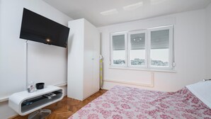 Quarto com cama, televisão na parede, janela com persianas e chão de madeira.