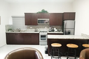 A modern kitchen with dark cabinets, a microwave, a refrigerator, a stove, and a granite countertop. There are three wooden stools and a brown leather chair.