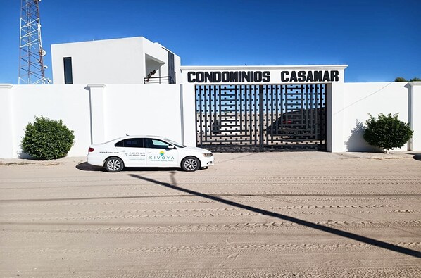 A white car parked in front of a gated entrance with the sign "CONDOMINIOS CASAMAR".