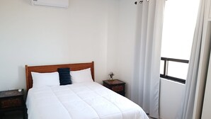 Un dormitorio con cama de madera, ropa de cama blanca, una mesita de noche con un adorno pequeño y una ventana con cortinas.