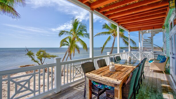 Terraza de madera con una mesa y sillas, con vista a una playa con palmeras y el océano.