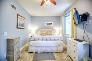 Une chambre à coucher comprenant un lit, des tables de chevet, une commode, un ventilateur de plafond, une fenêtre avec des rideaux et des œuvres d’art accrochées au mur.
