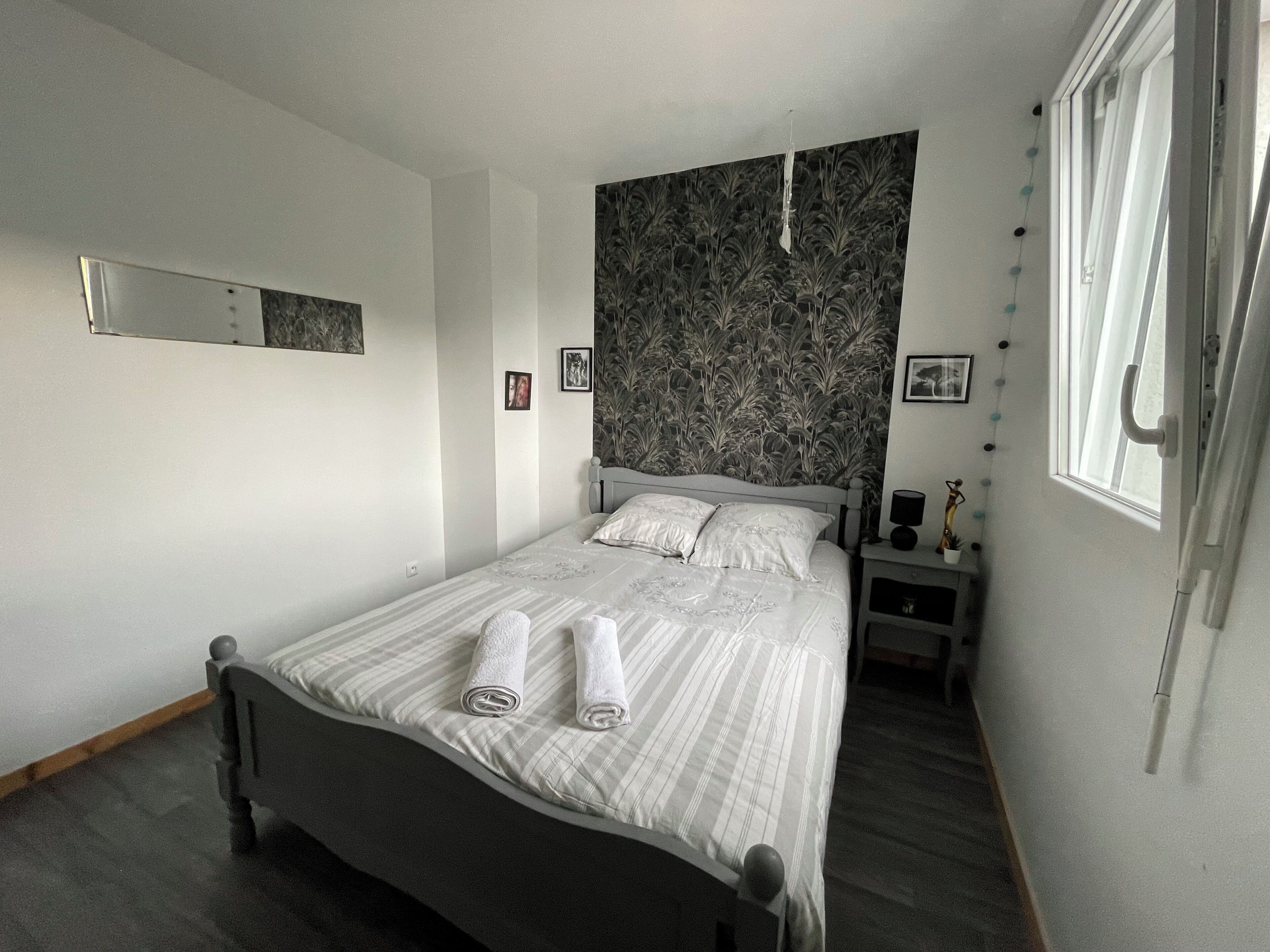 Una cama bien hecha con toallas enrolladas, una mesita de noche con lámpara, un espejo en la pared y una ventana con cortinas.