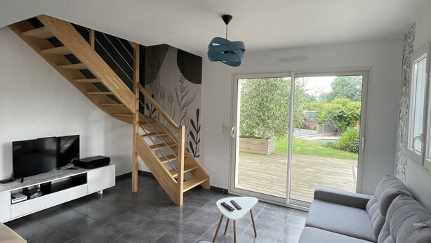 Una sala de estar moderna con un sofá gris, un televisor sobre un mueble blanco, una escalera de madera y vistas a un jardín a través de puertas de vidrio.
