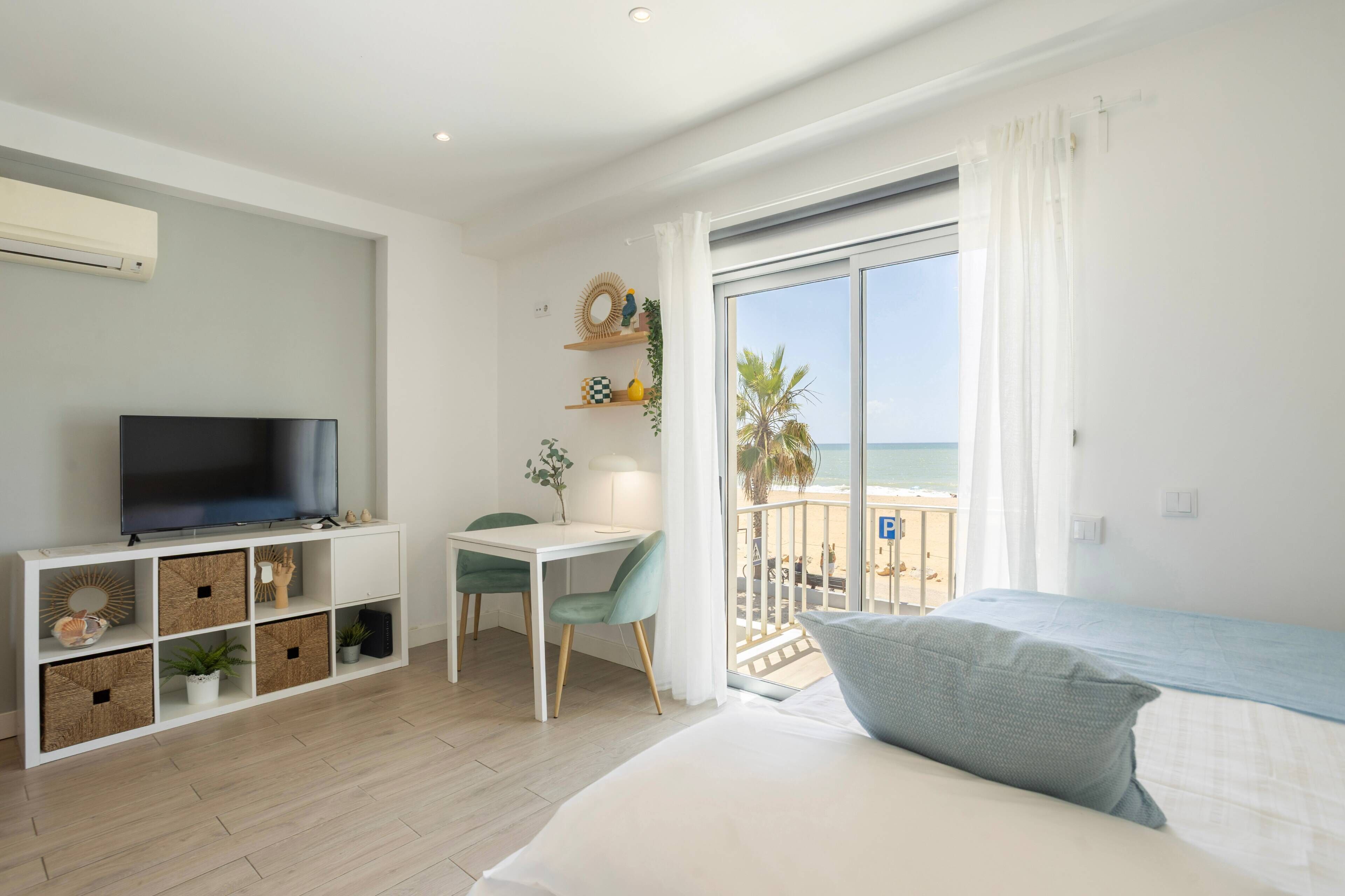A modern hotel room with a TV, a desk, a bed, and a balcony with a view of the beach.