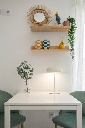 A white table with a lamp, two green chairs, a vase with green leaves, a wooden shelf with decorative items, and a wall-mounted mirror.