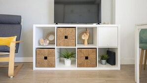 Ein Wohnzimmer mit einem Flachbildfernseher auf einer weißen Konsole, Weidenkörben, einer Topfpflanze und einem kleinen Lautsprecher.