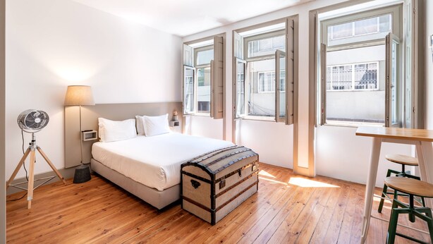 Un dormitorio con cama, suelo de madera, escritorio con silla y ventana con contraventanas abiertas.