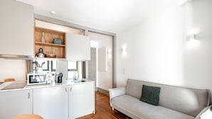 Une cuisine moderne avec des meubles blancs, un four à micro-ondes et une machine à café. On y trouve une table à manger en bois avec des chaises. Un canapé de couleur claire est placé contre le mur, et une porte donne accès à une autre pièce.