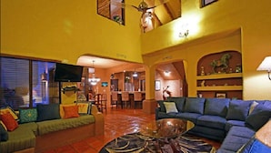 A spacious living room with a large sofa, a coffee table, and a TV. There is a dining area with chairs and a table in the background.