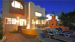 A modern building with a pool area featuring lounge chairs and a table, surrounded by a low wall and potted plants.