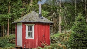 Eine kleine rote Holzhütte mit Schornstein, aus dem Rauch aufsteigt, umgeben von dichtem Wald.