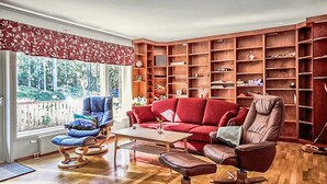 Ein Wohnzimmer mit einem roten Sofa, einem blauen Sessel und einem braunen Sessel. Eine Holzbibliothek ist mit Büchern und dekorativen Gegenständen gefüllt. Ein Fenster bietet Blick auf Bäume.