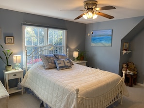 A bedroom with a bed, a ceiling fan, a window with a view of trees, a painting on the wall, and bedside tables with lamps and plants.