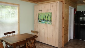 Een kamer met houten lambrisering, een eettafel met stoelen, een schilderij van een boslandschap en een zwarte koelkast.