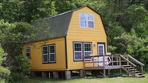 Een geel houten huis met een klein terras en een trap, omringd door bomen.