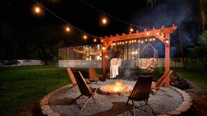 Ein gemütlicher Außenbereich mit Feuerstelle, Holzhöhlen und einer Hängematte unter einer Pergola mit Lichterkette.