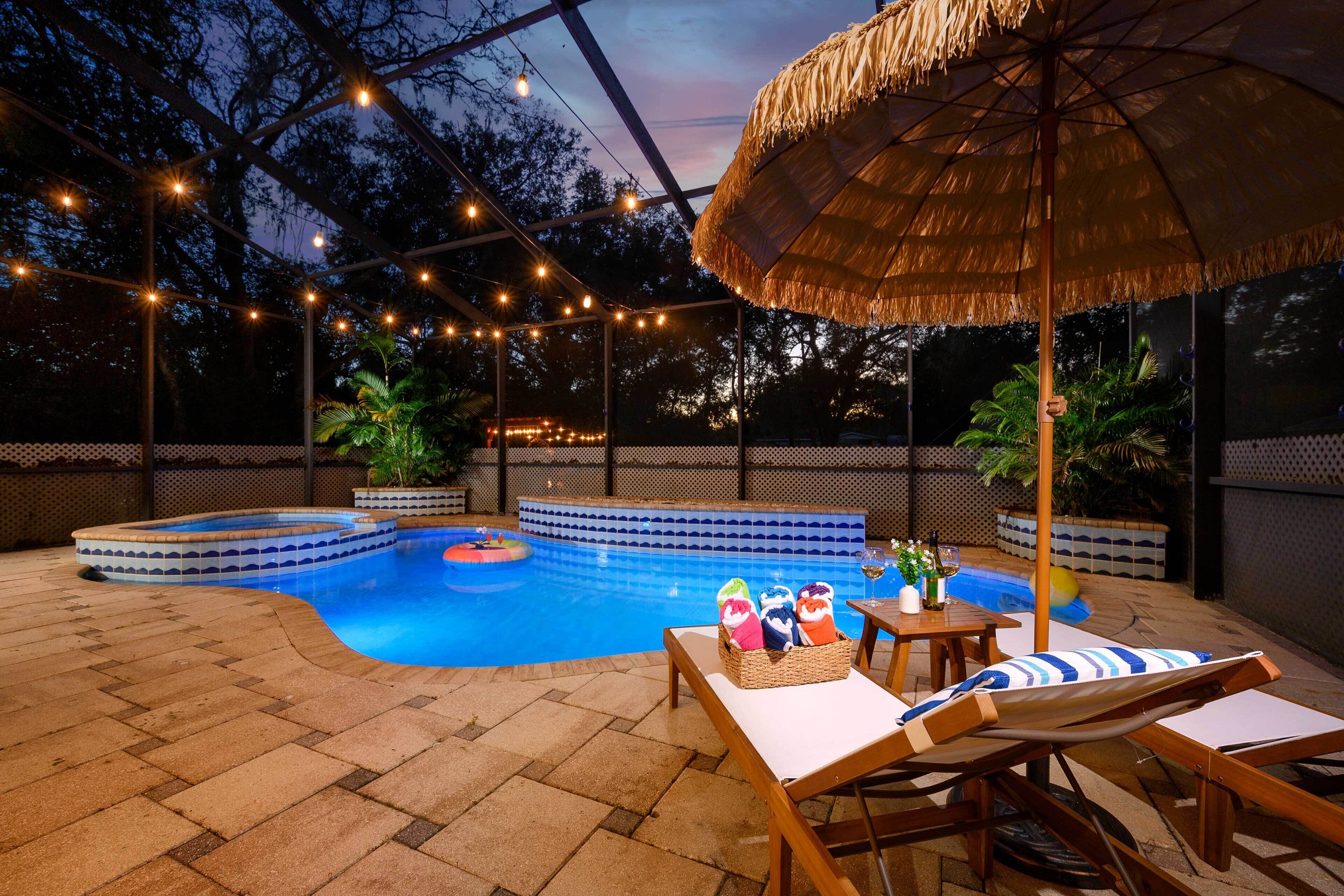 An outdoor swimming pool area with lounge chairs, a thatched umbrella, and string lights.