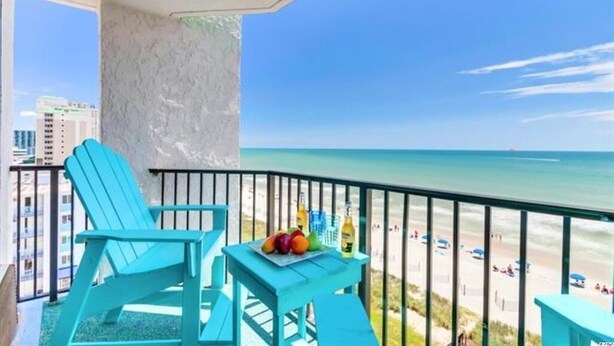 A balcony with turquoise chairs and a table with fruit and drinks, overlooking a beach with people and buildings in the background. - Newly Renovated Beachfront Oasis. (Myrtle Beach)