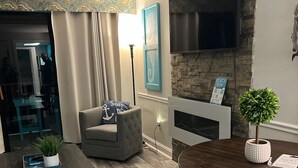 A living room with a grey sofa, a wooden coffee table, a dining table with four chairs, a flat-screen TV mounted on a stone wall, and a small potted plant. - Newly Renovated Beachfront Oasis. (Myrtle Beach)