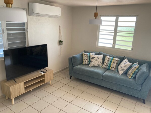 A living room with a TV on a wooden stand, a blue sofa with patterned cushions, a hanging light, and a window with blinds. - Walking distance to Boqueron, private pool, full A/C, sleeps 10 (Boquerón)
