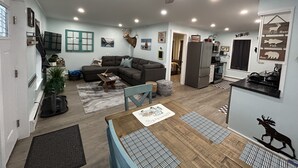 En rummelig opholdsstue med en stor sofa, et spisebord med servietter, og et køkkenområde med et køleskab og en mikroovn.