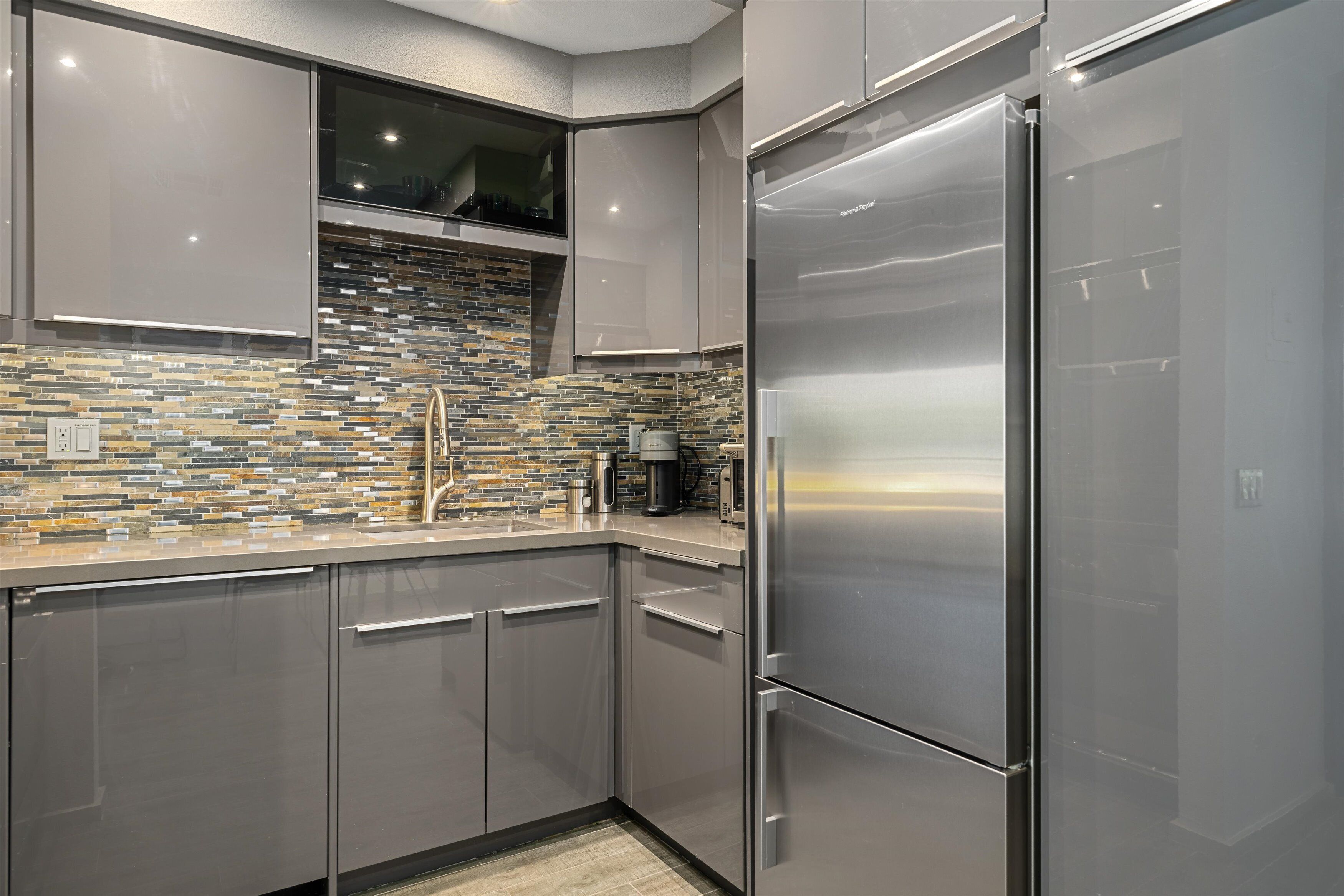 A modern kitchen with a stainless steel refrigerator, gray cabinets, and a mosaic tile backsplash.