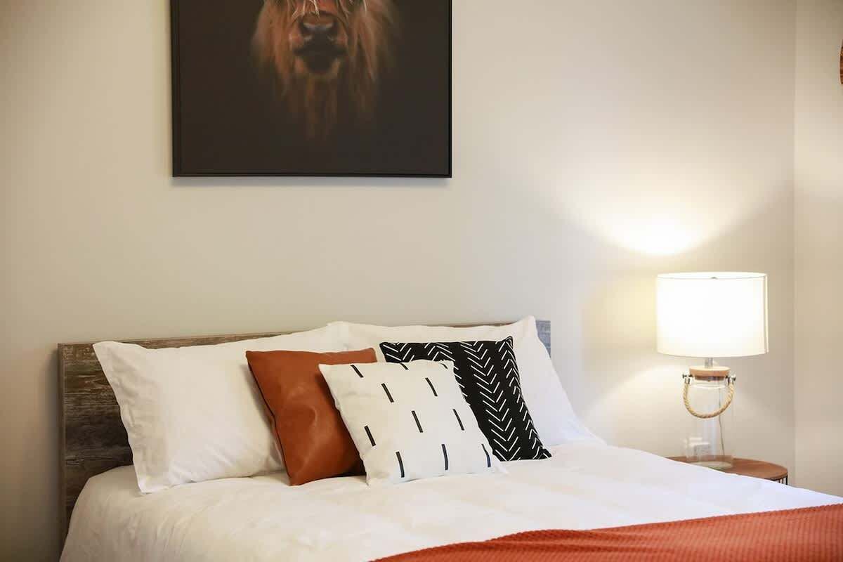 A neatly made bed with white bedding and multiple pillows, a wall-mounted artwork above the bed, and a bedside lamp on a wooden nightstand.